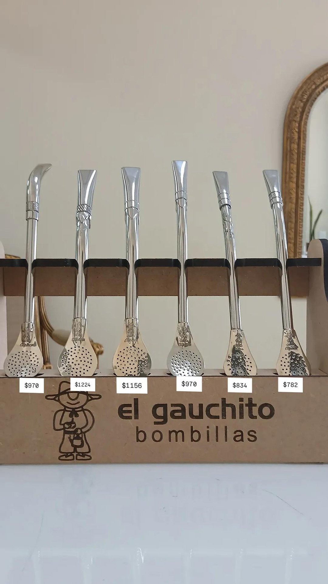 Exhibidor de madera con seis bombillas de alpaca marca El Gauchito para mate, con filtro tipo cuchara y diferentes diseños labrados, con precios desde $782 a $1224.